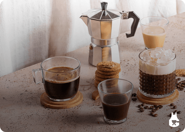 Coffee setup with moka pot, espresso, iced coffee, and cookies on a stone surface — welcoming beginner scene.